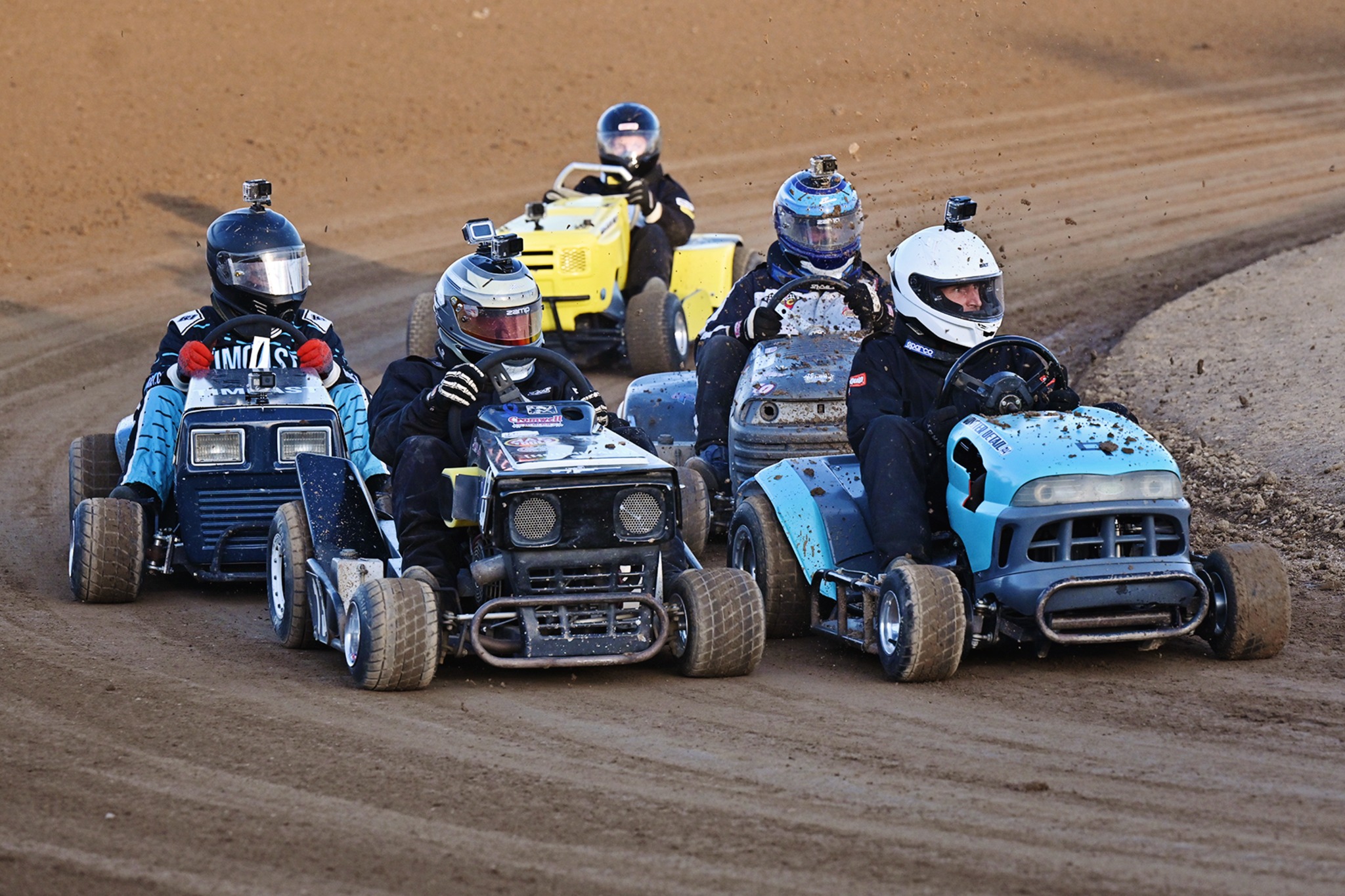 A tight pack of five helmeted lawnmower racers rounding a dirt-track corner